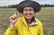 Anna Tulley in a farm field squinching two of her fingers together