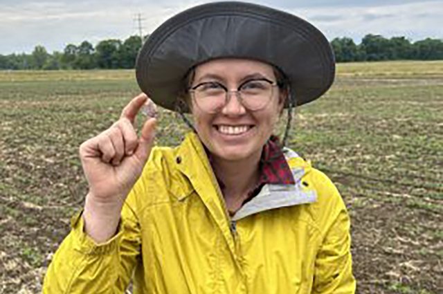Anna Tulley in a farm field squinching two of her fingers together