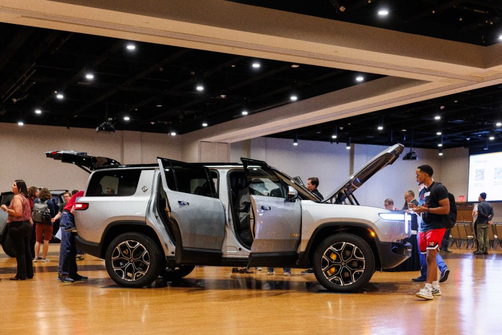 A Rivian R1S SUV is parked in the Brown Ballroom with all of its doors open, as people look at the vehicle