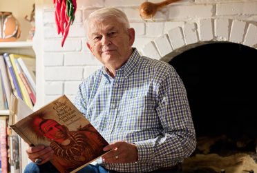 photograph of Dr. Edmunds holding a book titled "The George Catlin Book of American Indians" sitting near a fireplace