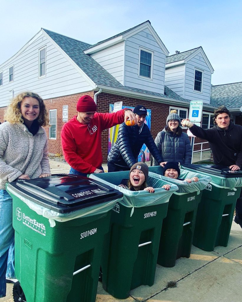 Three students sitting inside three green compost bins with five students behind them. One student is holding a small pumpkin over the bins to compost.