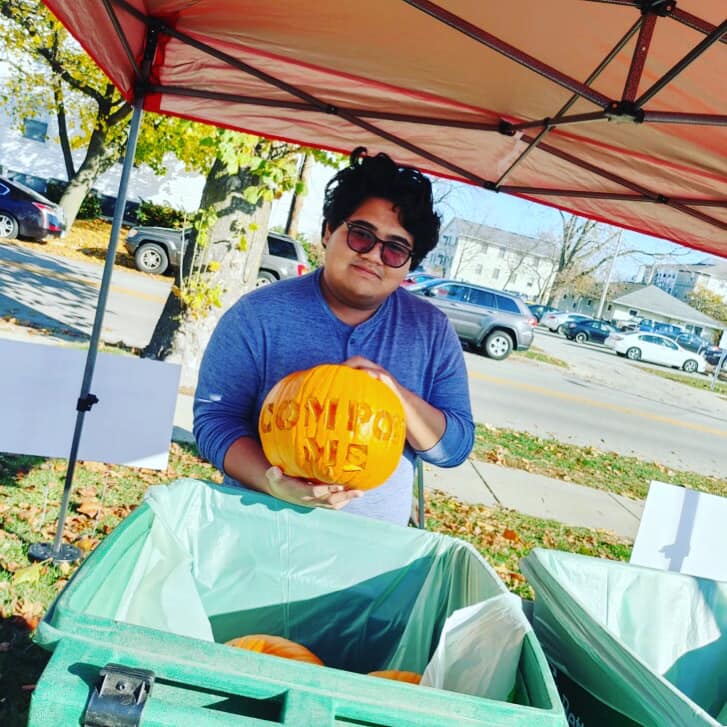 Student holding a pumpkin carved with the world "Compost Me" behind a green compost bin.
