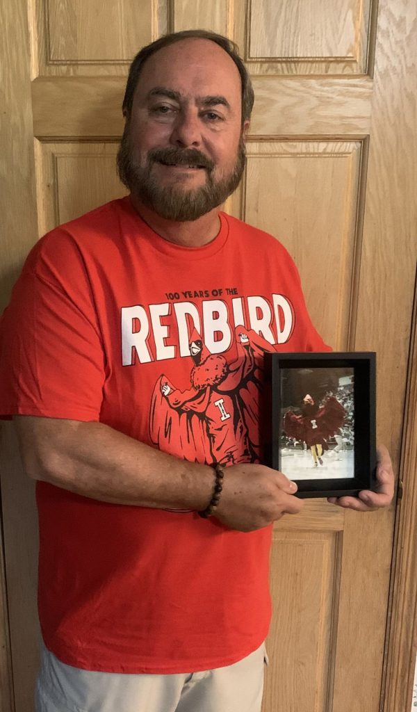 Man standing in front of door holding framed photo of Redbird mascot, wearing T-shirt featuring the same image