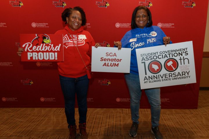 two individuals holding signs reading "Redbird proud," "SGA senator alum" and "student government association's 100th anniversary" pose in front of a backdrop at student government anniversary event