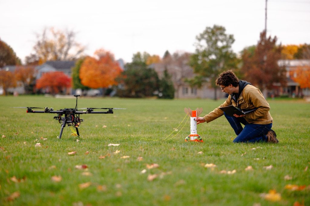 A person holding a laptop prepares a rocket, which is attached to a drone on the ground.