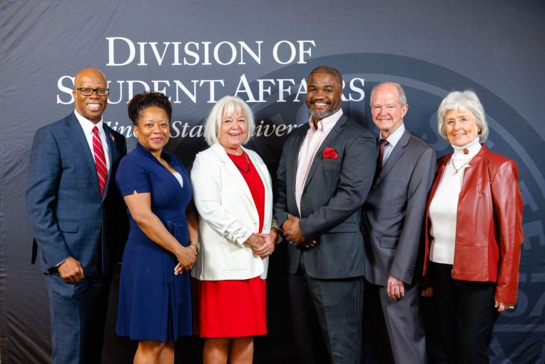 Six people standing and smiling in front of the Division of Student Affairs logo