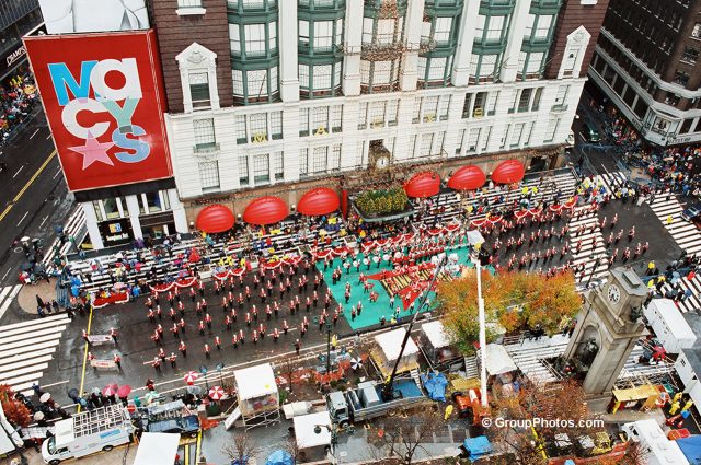 Aerial view of a marching band performing on a street in front of a tall building with a "Macy's" sign on the corner.