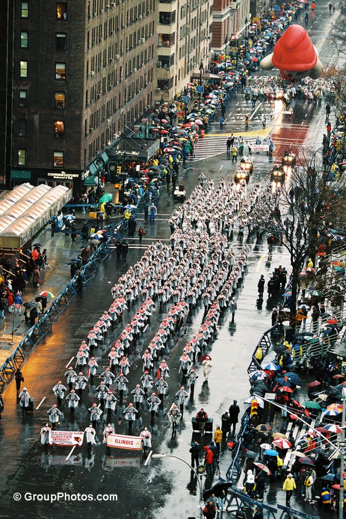 Aerial view of a marching band marching down a street, surrounded by tall buildings.