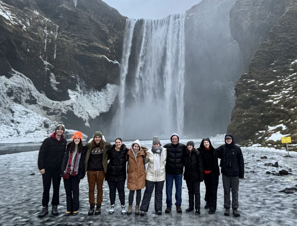 A group of 10 students stand on a glacier in front of a waterfall.