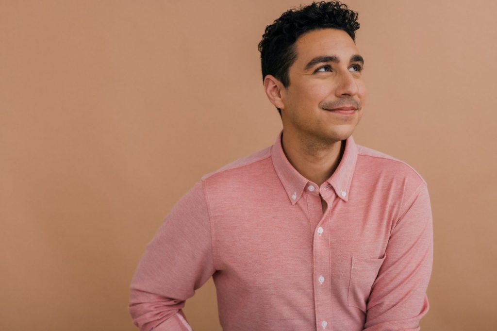 A photo of Joey Contreras against a dark tan background. He is a young man with dark hair, and he is wearing a pink button down.