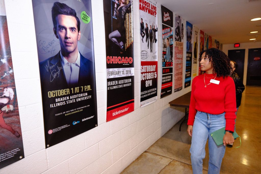 Angelina Ruiz looks up at a line of posters featuring various previous Illinois State guest performers; she stand directly across from a poster of magician Michael Carbonaro. 