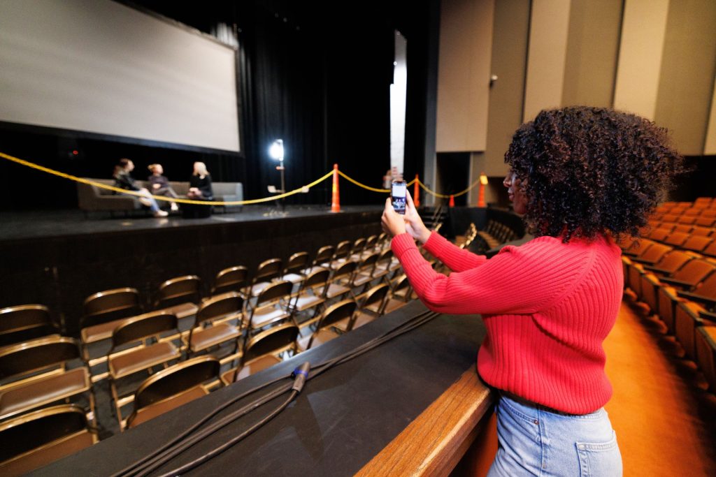 Angelina Ruiz stands in the seating area of Braden Auditorium facing the stage and holding her phone to test camera angles. 