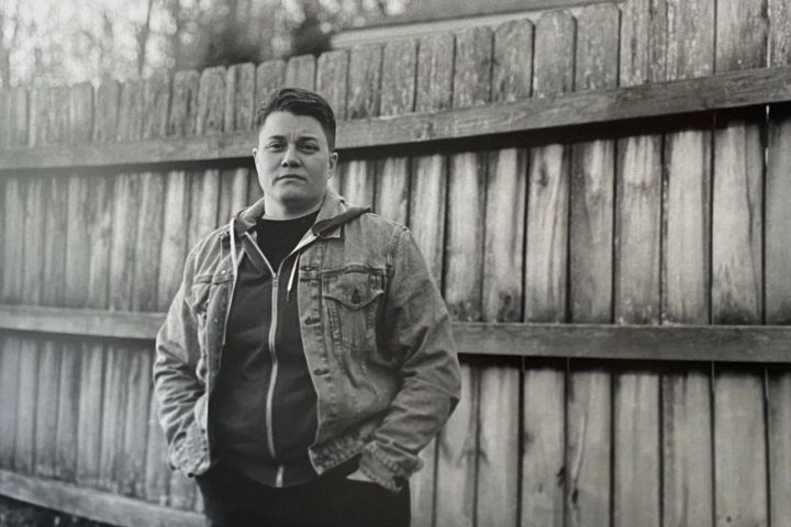 A black and white photo of a white nonbinary person, Dr. Derek Sparby, in front of a fence.