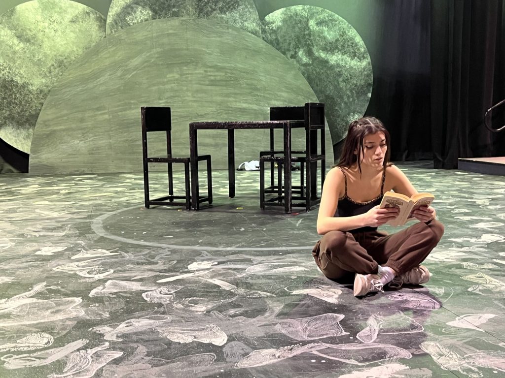 Bryce Henderson as Arugula, sitting cross-legged on a mostly empty, black and white stage. She is wearing brown pants and a black tank top. In the background is a black table and chairs and a collage of moons and planets.
