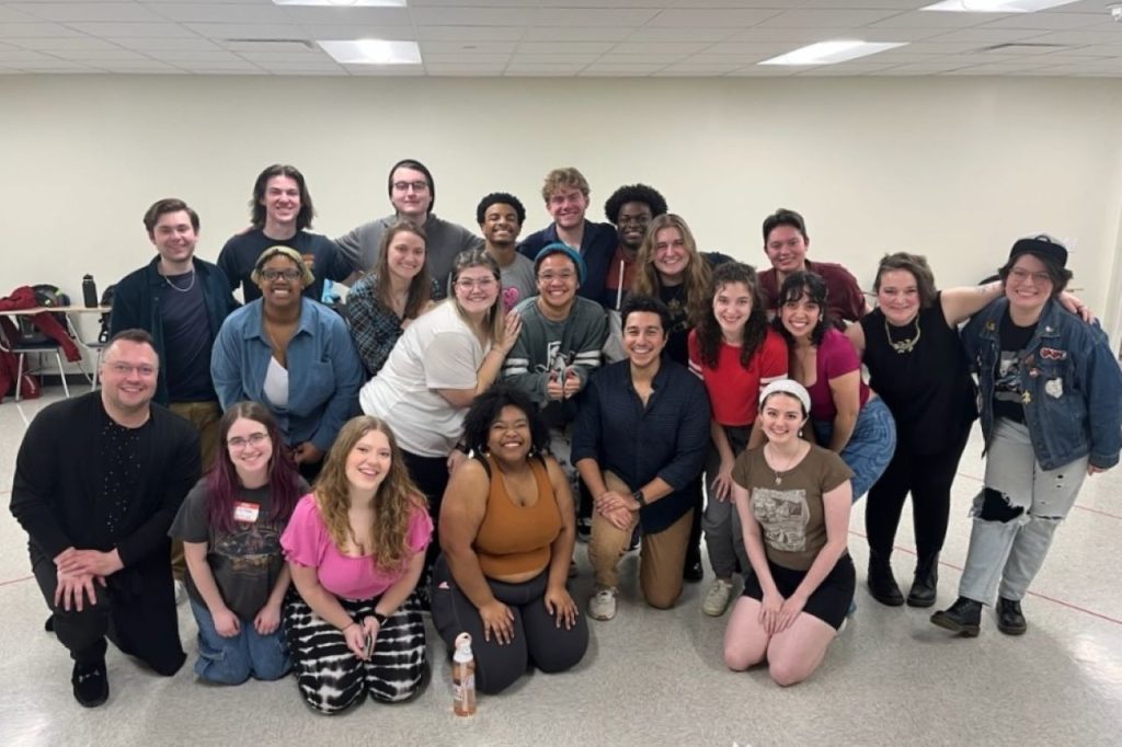 A casual group photo of the cast and creative team with Joey Contreras in their rehearsal space. Contreras is kneeling at the front and center of the group.