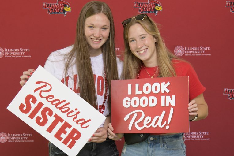 two girls hold signs. One says Redbird sister. Other sign says I look good in red