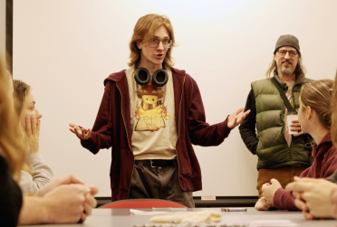 A student, standing with headphones around their neck, presents a game