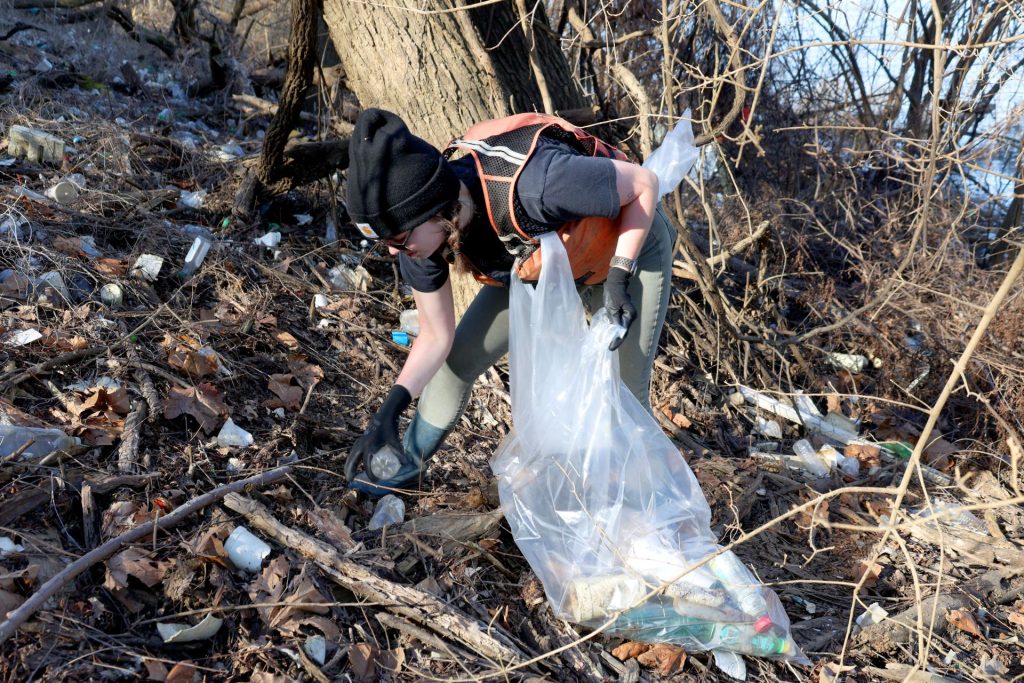 person picks up trash along the shoreline holding a trash bag and wearing a life jacket