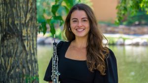 Headshot for Jillian Kouzel holding her oboe. The background is an outside scene with trees, bushes, and water.