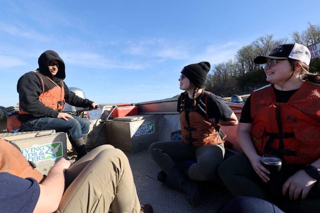 three people sitting on a boat with one steering and all wearing life jackets 