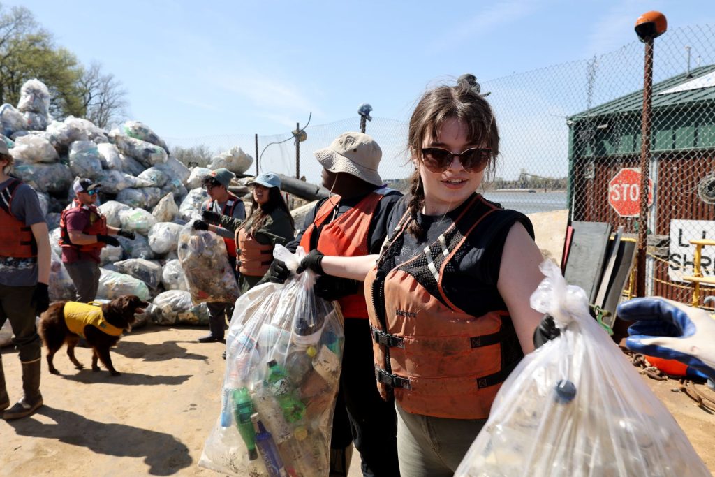 Person standing in a line of people holding two trash bags with a pile of trash in the background.
