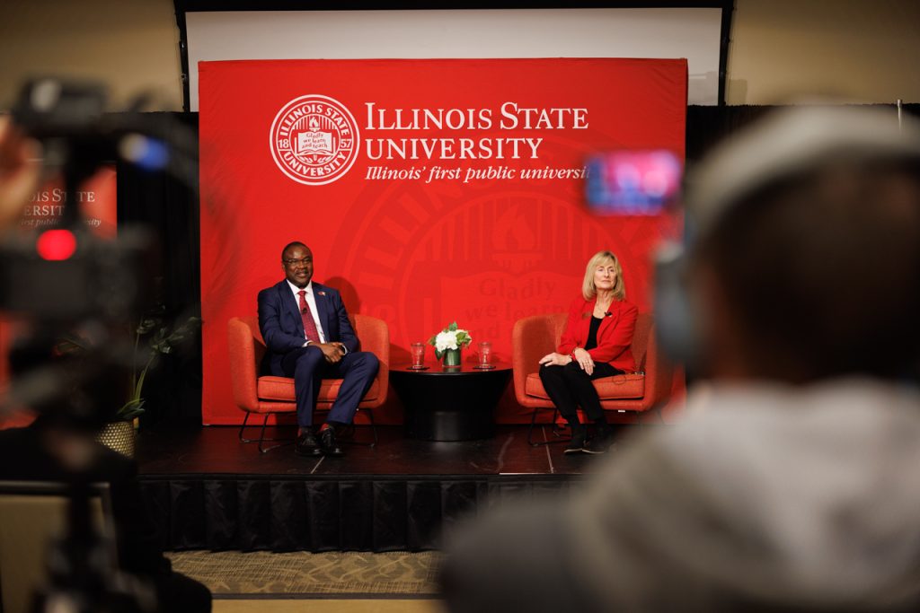President Aondover Tarhule and Dr. Kathy Bohn sit in front of a red backdrop with "Illinois State University" and the seal, as news cameras look on