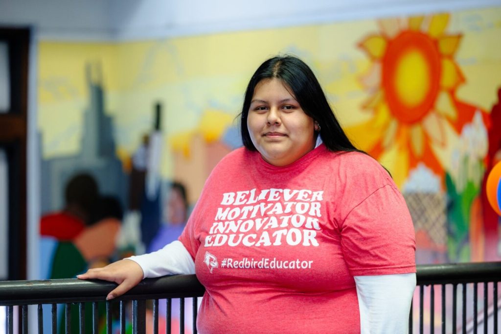 Student Yehiri Gonzalez stands wearing a red t-shirt that says "Believer, motivator, innovator, educator," in front of mural.