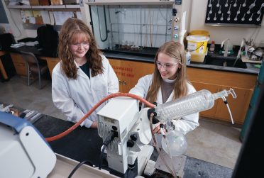 Two students in a chemistry lab with equipment and wearing safety goggles