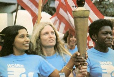 Three women attending the National Women's Conference in 1977. They are wearing matching shirts and holding onto a torch.