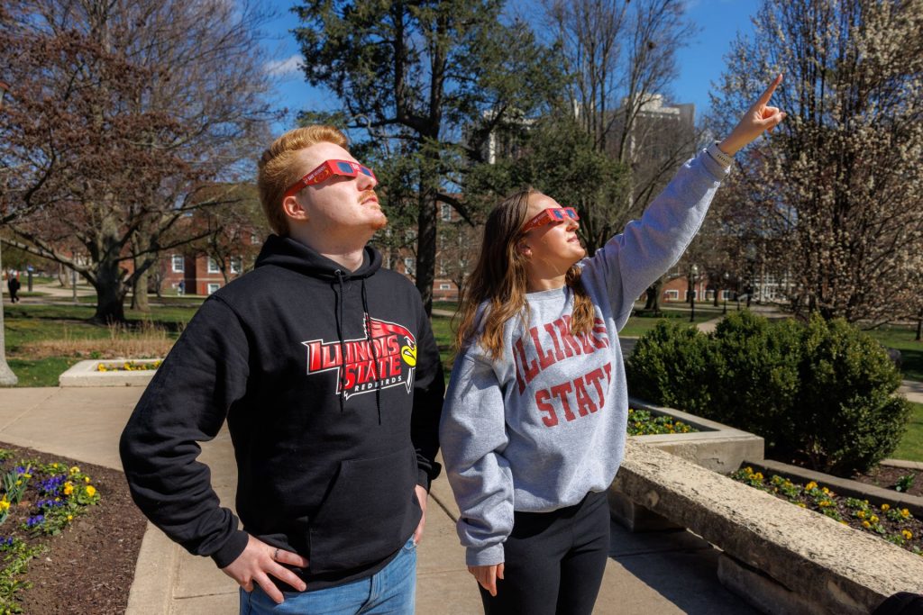two students wearing solar eclipse glasses on campus looking up, one pointing, one with hands on hips