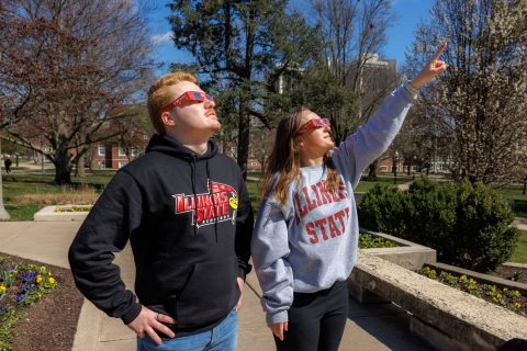 two students wearing solar eclipse glasses on campus looking up, one pointing, one with hands on hips
