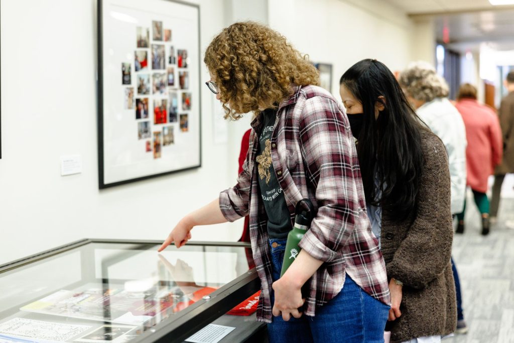 A student points out a the WGSS artefact encased in a display table to a friend with a collage of photos on the wall in front of them. 