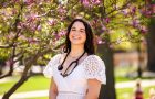 Olivia Fedorko stands beneath a tree on the quad wearing a white dress with a stethoscope draped around her neck