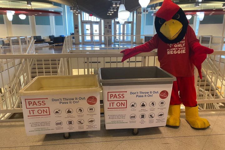 Reggie Redbird mascot stands next to two Pass It On bins.