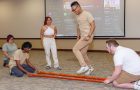 Two people sit on floor holding two poles while one person hops in between to perform Tinikling while two other people watch.