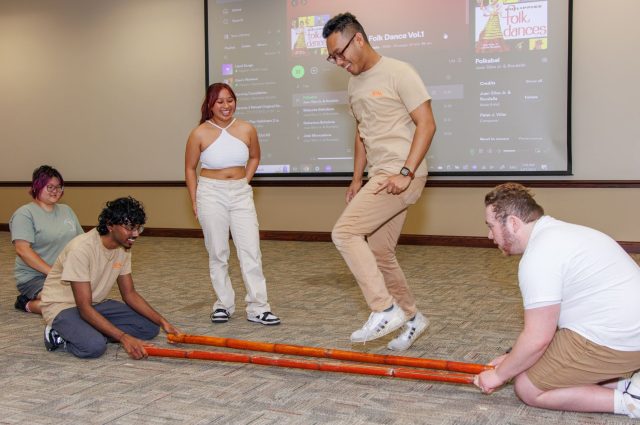 Two people sit on floor aholding two bamboo poles while one person hops in between to perform Tinikling while two other people watch.