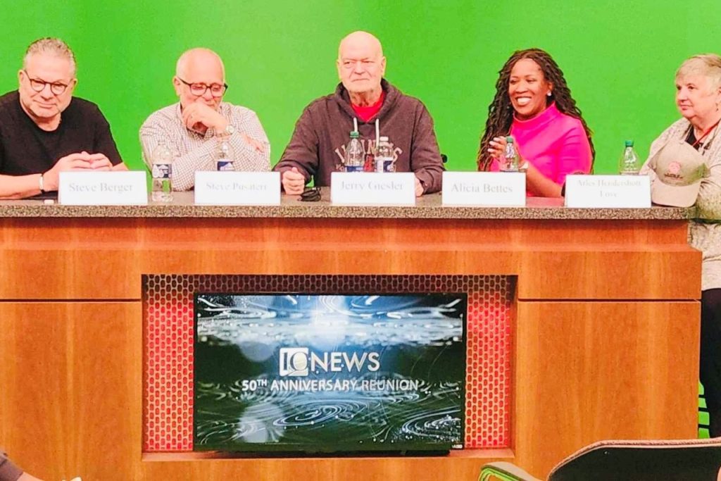 five people sitting at a news desk with a green screen behind them.