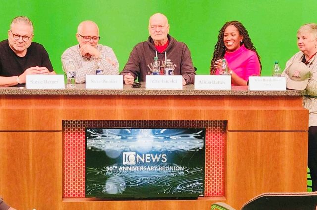 five people sitting at a news desk with a green screen behind them.