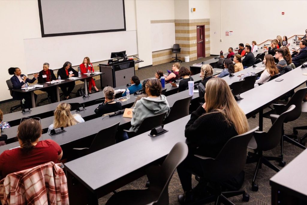 A panel of four women speaking to a large group of people in a lecture hall.