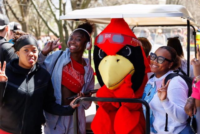 Three students pose with Reggie Redbird, with Eclipse glasses on his forehead