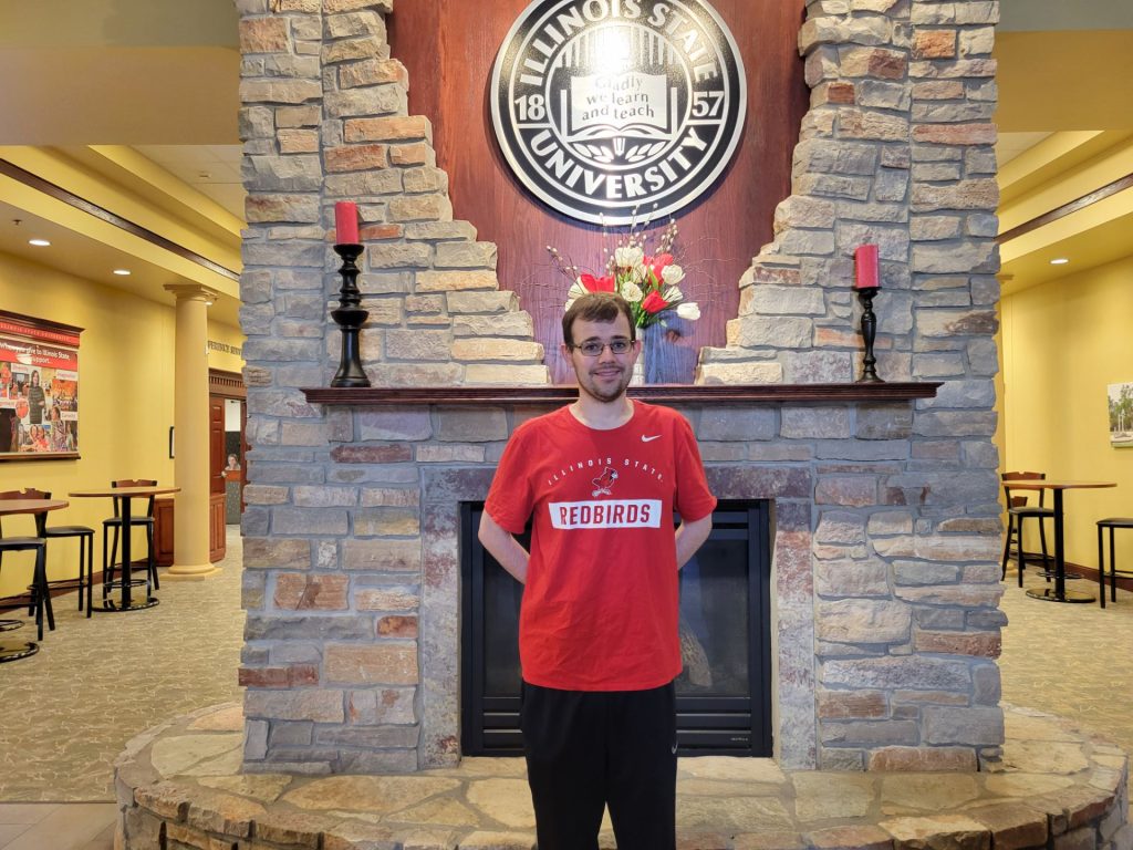 Avery Birnbaum wears a red t-shirt and stands in front of a fireplace with an Illinois State logo above and behind him on the wall.