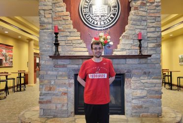 Avery Birnbaum wears a red t-shirt and stands in front of a fireplace with an Illinois State logo above and behind him on the wall.