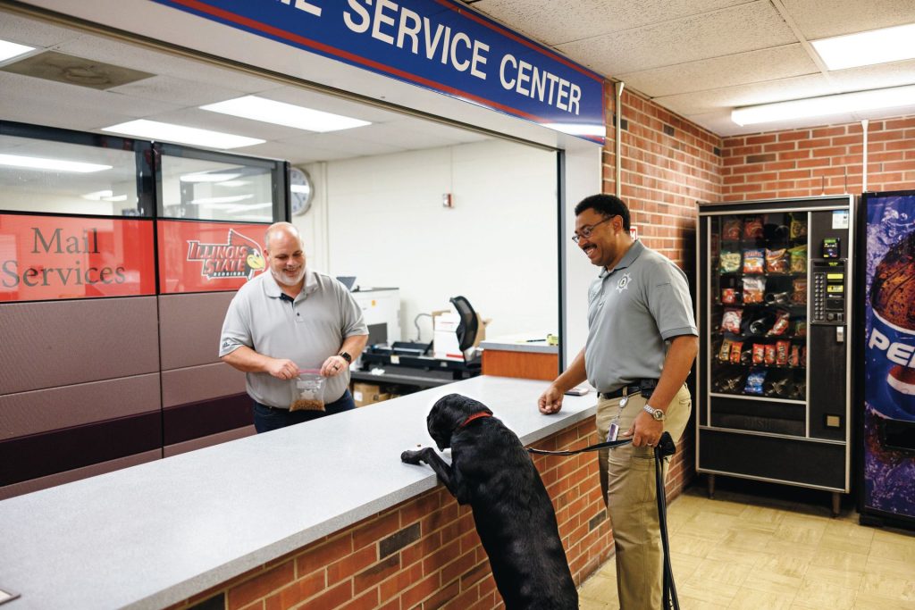 Mike Gardner holds Pawficcer Sage by her leash. Sage's stands on her hind legs with her front paws on the Mailing Services counter as Shannon Covey delivers a treat. 