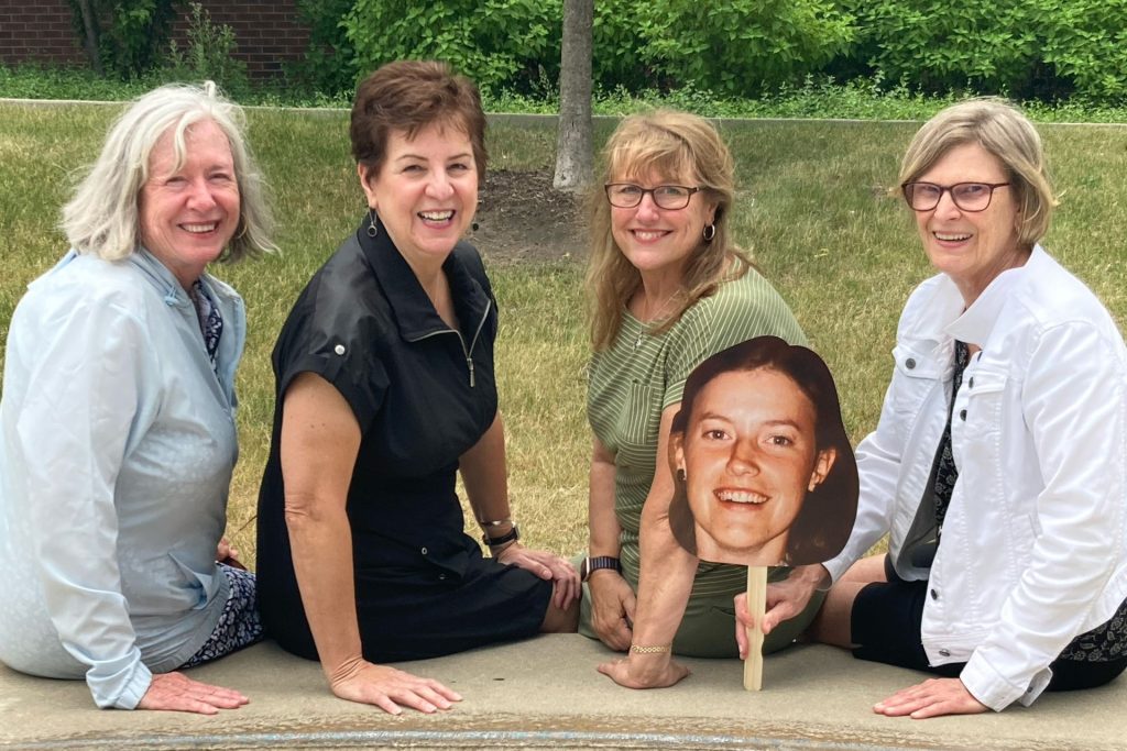 Four women seated on a bench, one holding a picture of a late classmate