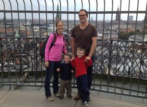 tqo adulst and two children stand on a balcony with a wrought iron fence, a view of Denmark in the background 