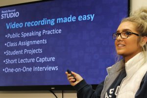 Female student using pointer with presentation with following text: Ready Record Studio Video Recording Made Easy Public Speaking Practice Class Assignments Student Projects Short Lecture Capture One-on-One Interviews