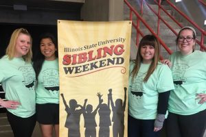 Four women smile in front of Sibling Weekend sign